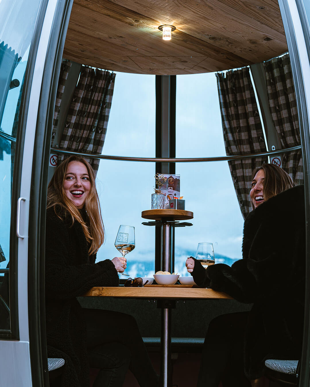 Zwei Frauen sitzen in einer Gondel mit einem Glas Wein und einer Schüssel Chips - Alpres Restaurant Après Ski